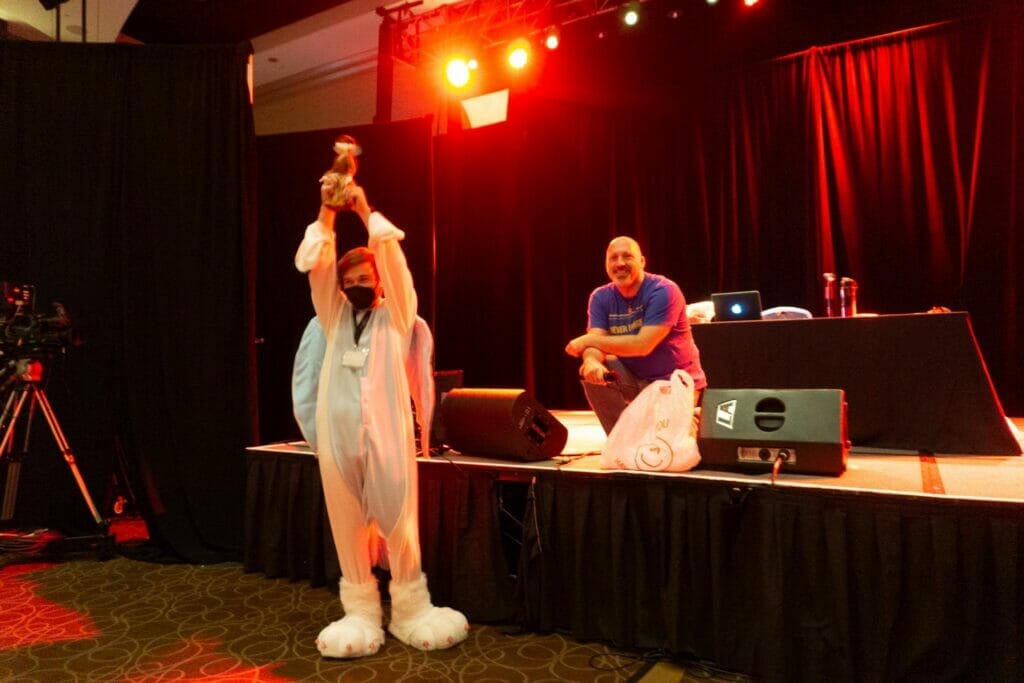 Photo of a man in a costume holding up a plush dog triumphantly. A man in a blue shirt is crouched on a stage behind him, smirking.