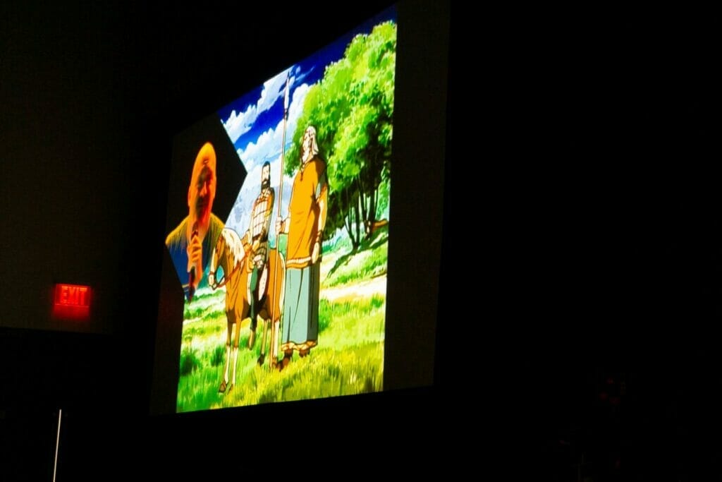 Photo of a cartoon image of a giant man next to a horse. We call him Wandering Perspective Man. Anyway, there's also a feed on the left side featuring a bald man in a blue shirt, who's talking into a microphone.