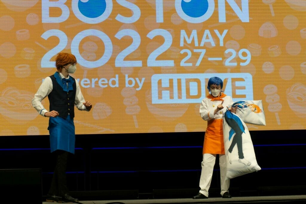 Photo from Anime Boston, which features a man with orange hair in dressy casual talking with a blue-haired woman in a chef's jacket. The woman is holding a dakimakura of an anime character.