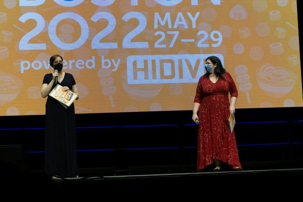 Photo from Anime Boston 2022's opening ceremonies. Kristin Leiding and Jenna Leary address the crowd in front of a screen with the text "Anime Boston 2022" projecting on it.