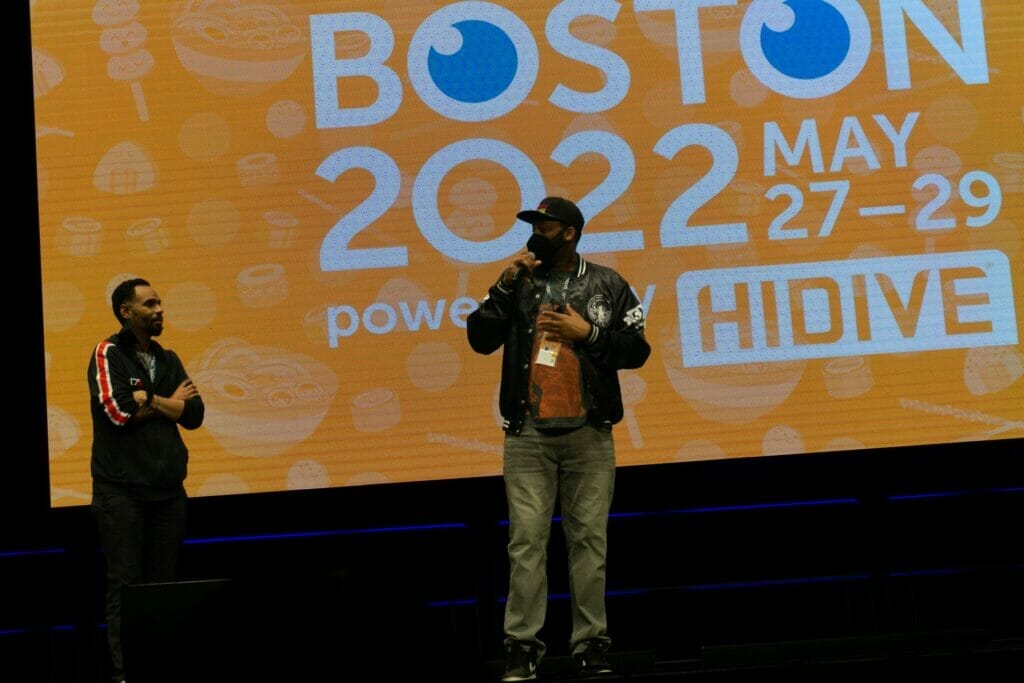 Photo from Anime Boston 2022's opening ceremonies. Rapper EyeQ address the crowd in front of a screen with the text "Anime Boston 2022" projecting on it.