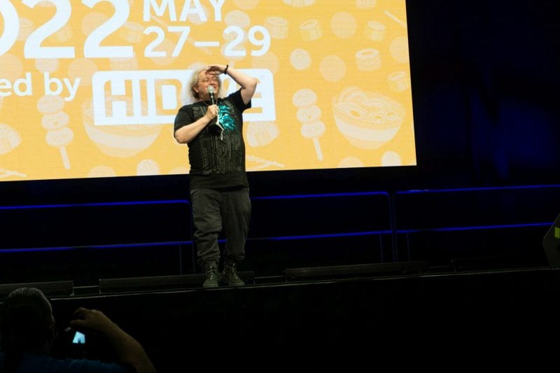 Photo from Anime Boston 2022's opening ceremonies. Greg Ayres gazes out at the audience in front of a screen with the text "Anime Boston 2022" projecting on it.