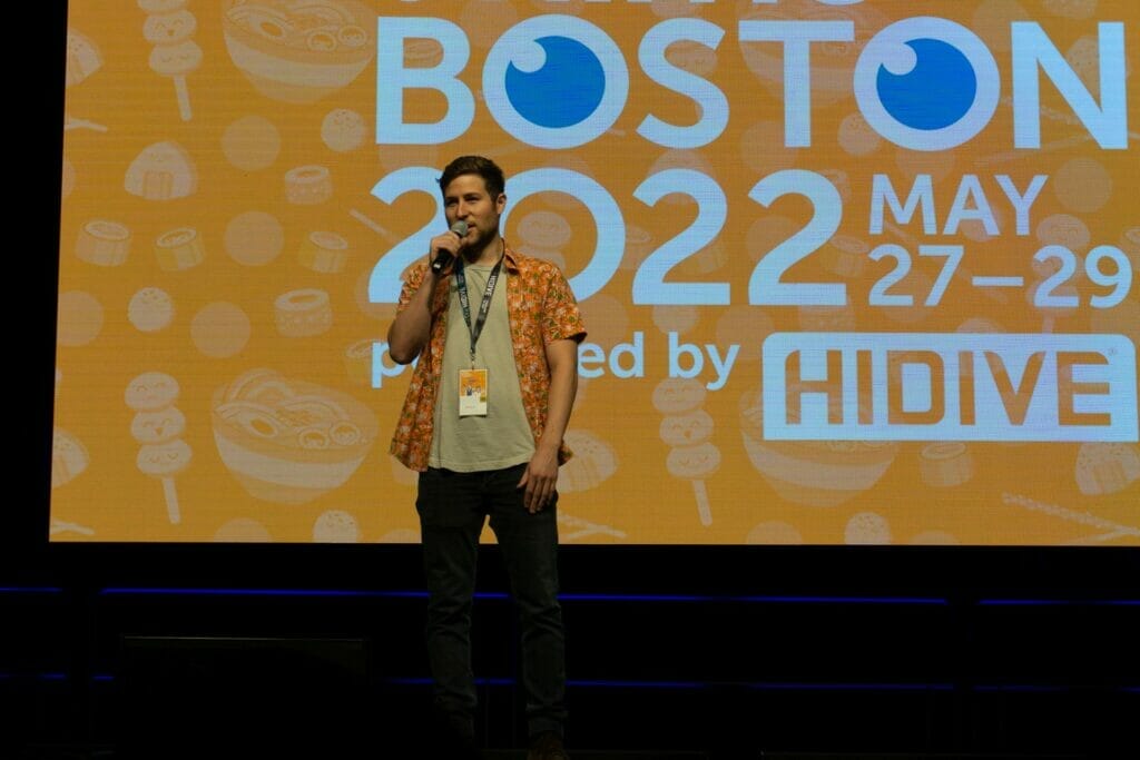 Photo from Anime Boston 2022's opening ceremonies. Griffin Burns address the crowd in front of a screen with the text "Anime Boston 2022" projecting on it.