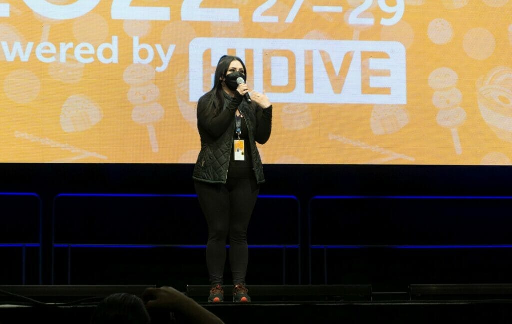 Photo from Anime Boston 2022's opening ceremonies. Lauren Landa address the crowd in front of a screen with the text "Anime Boston 2022" projecting on it.