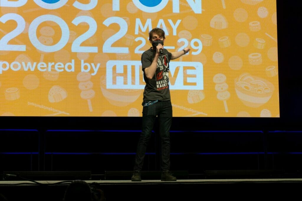 Photo from Anime Boston 2022's opening ceremonies. Matt Shipman address the crowd in front of a screen with the text "Anime Boston 2022" projecting on it.