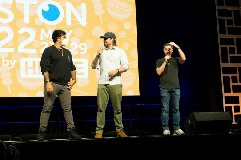 Photo from Anime Boston 2022's opening ceremonies. Max Mittelman, Ray Chase, and Robbie Daymond greet the crowd in front of a screen with the text "Anime Boston 2022" projecting on it.
