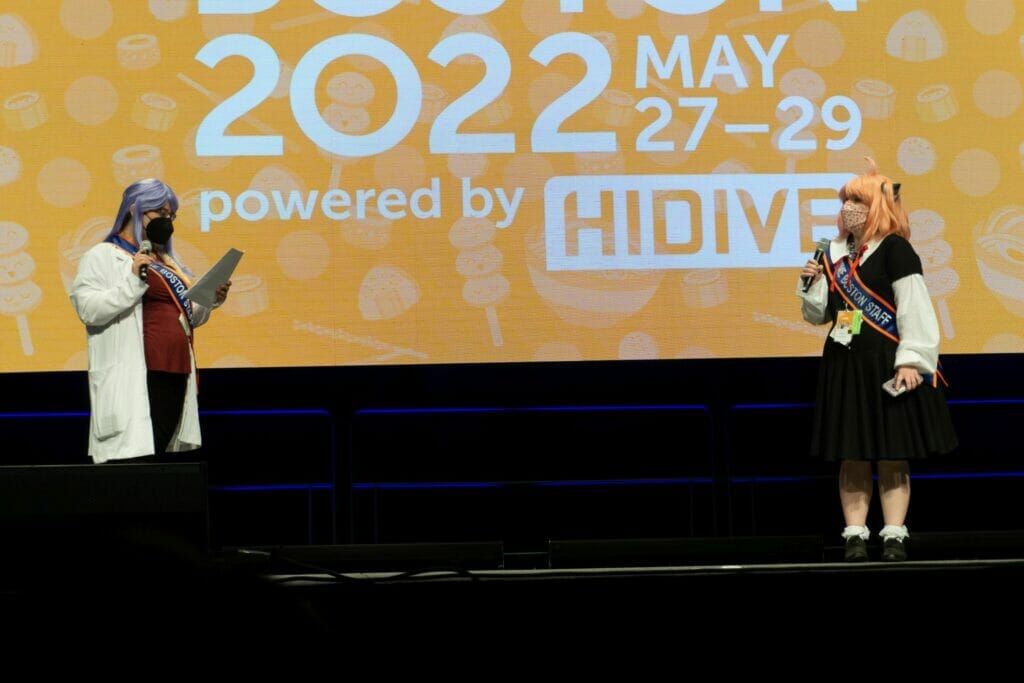 Photo from Anime Boston 2022's opening ceremonies. An individual dressed as a doctor and a person dressed as Anya from Spy x Family address the audience.