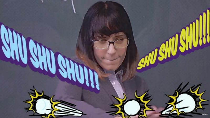 A brown-haired woman wearing a suit and glasses launches silver balls at the camera. Text reads "Shu Shu Shu!!! Shu Shu Shu!!!"