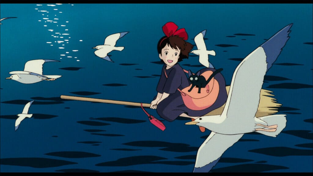 Screenshot from Kiki's Delivery Service, which features a young girl in a blue dress, wearing a red hair ribbon. She's riding a broom over the sea, aid a flock of gulls.