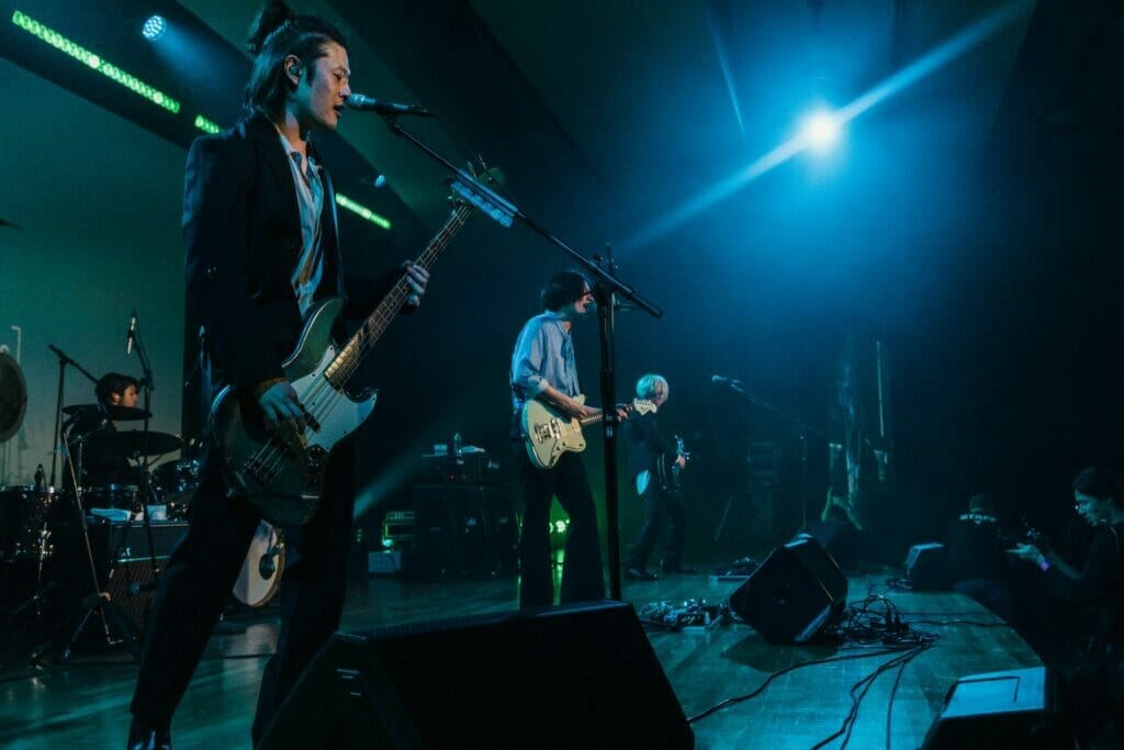 Ba thành viên của nhóm nhạc rock [Alexandros] đứng trên sân khấu trong một buổi hòa nhạc, cầm đàn guitar.  Họ đang hát vào micro đứng.