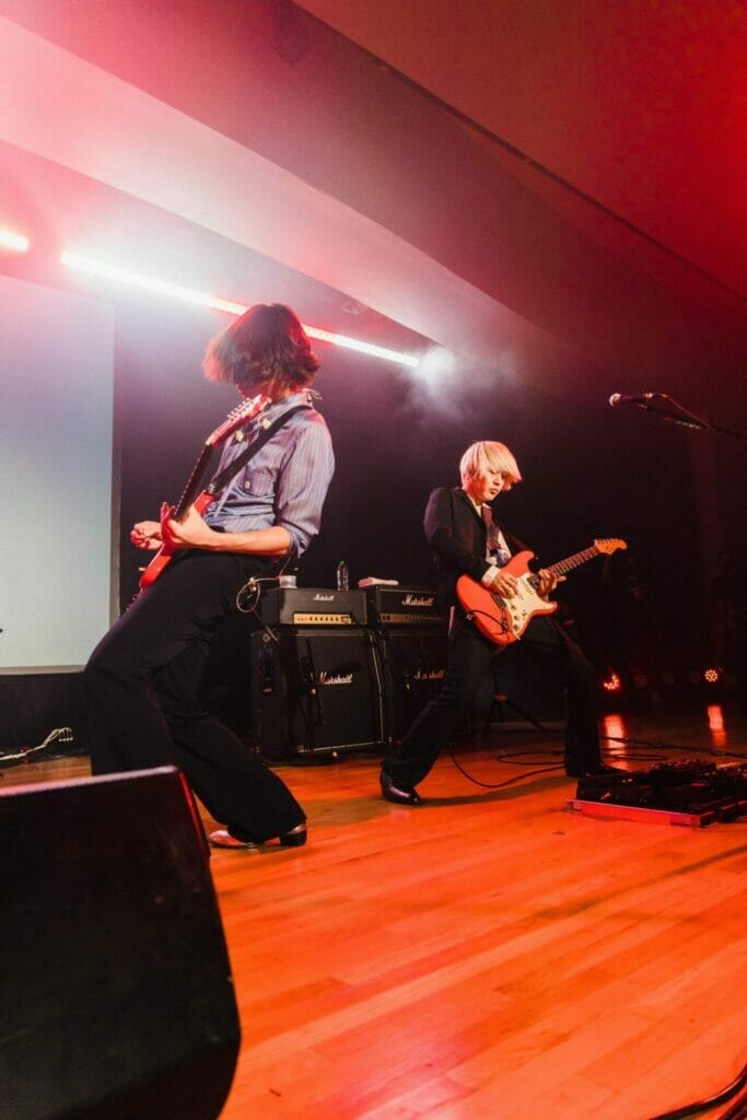 Photograph of two members of [Alexandros] playing guitar back-to-back on stage.