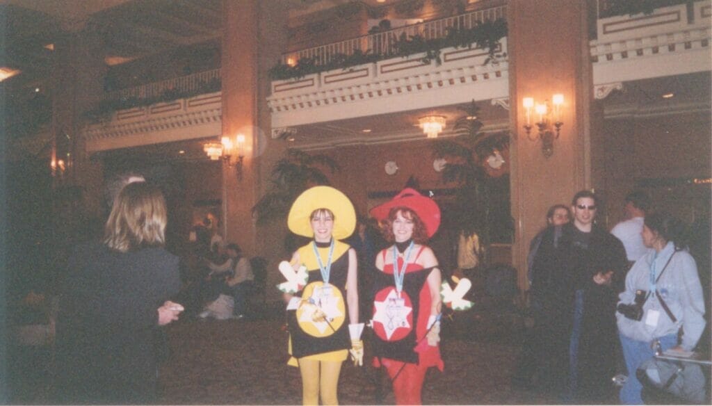 Photograph of two women dressed as colorful witch costumes, posing in a hotel lobby. They're flanked by people passing by, who look on curiously.