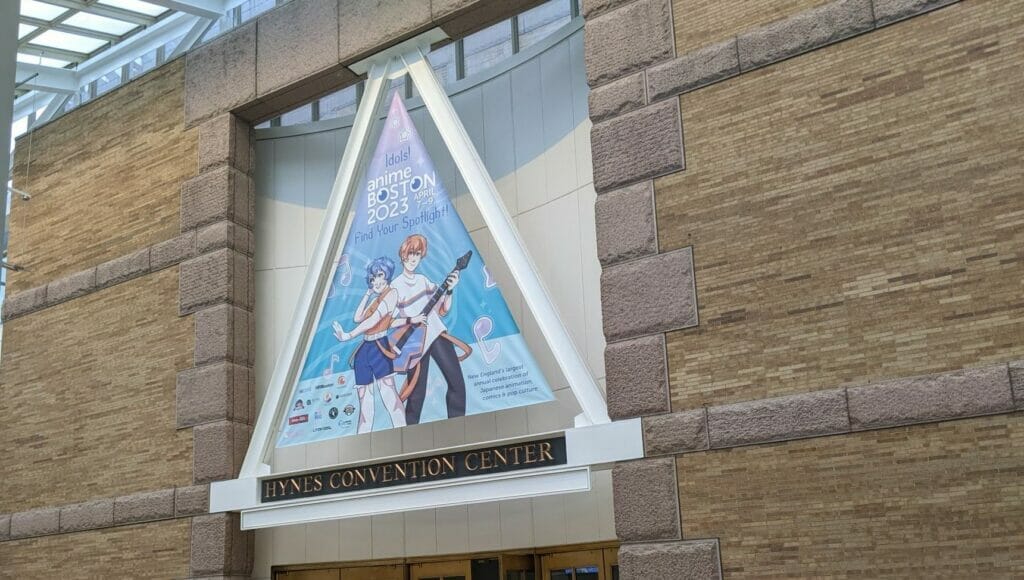 Photo from Anime Boston 2023 that depicts the event's sign, which features an orange-haired man and a blue-haired woman dressed in casual clothes. The man is holding a blue-and-orange guitar.Text: Idols! Find Your Spotlight! Anime Boston 2023 April 7 - 9