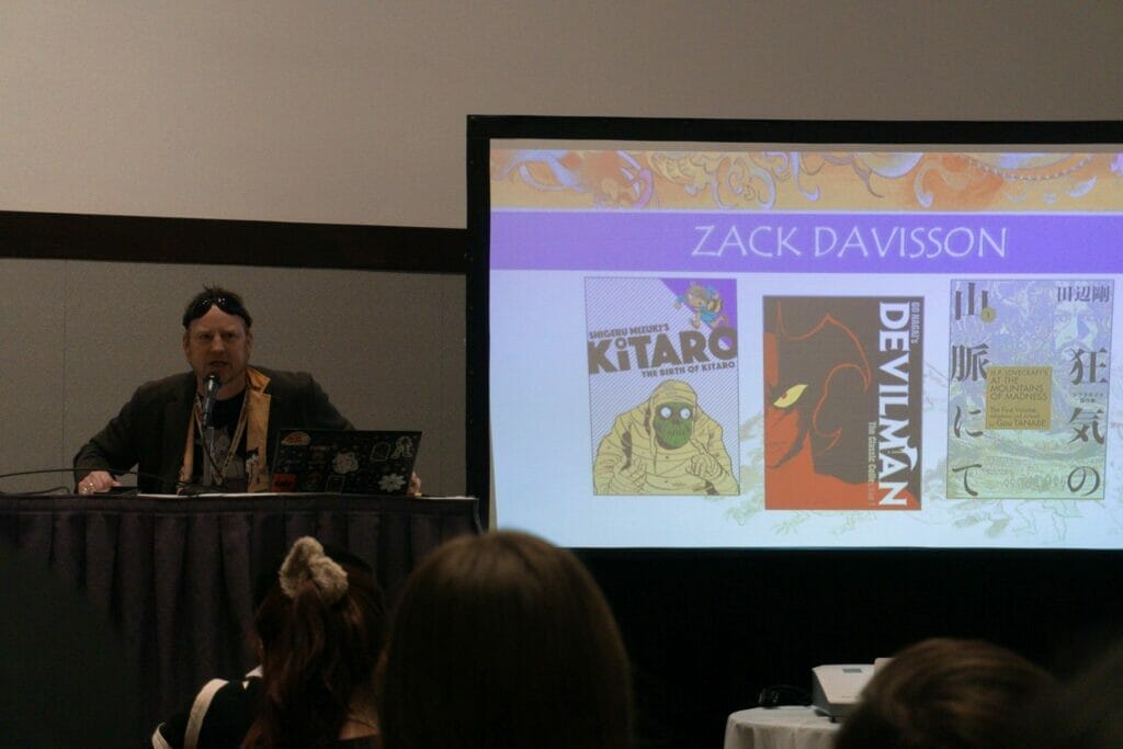Zack Davisson, a man with short hair and a light beard, sitting next to a PowerPoint slide with his name on it. He's wearing a sport jacket and T-shirt, and a pair of goggles rests at his hairline.