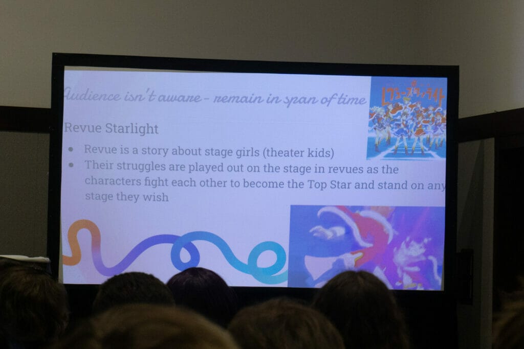 Photo of a PowerPoint slide with text. Text: "Audience isn't aware - remain in span of time" "Revue Starlight" "Revue Starlight is a story about stage girls (theater kids)" "Their struggles are played out on the stage in revues as the characters fight each other to become the Top Star and stand on any stage they wish"