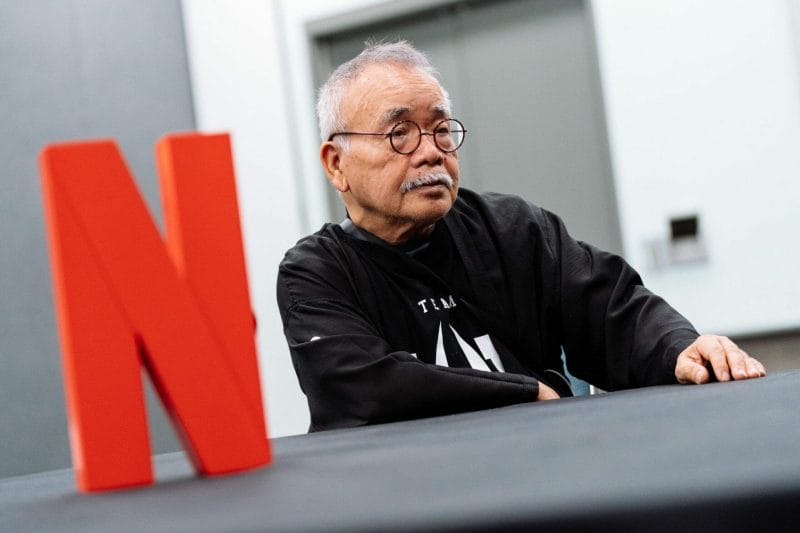 Anime producer Masao Maruyama sits beside a large, red Netflix "N". He's waring a black hoodie and a Pluto T-shirt.