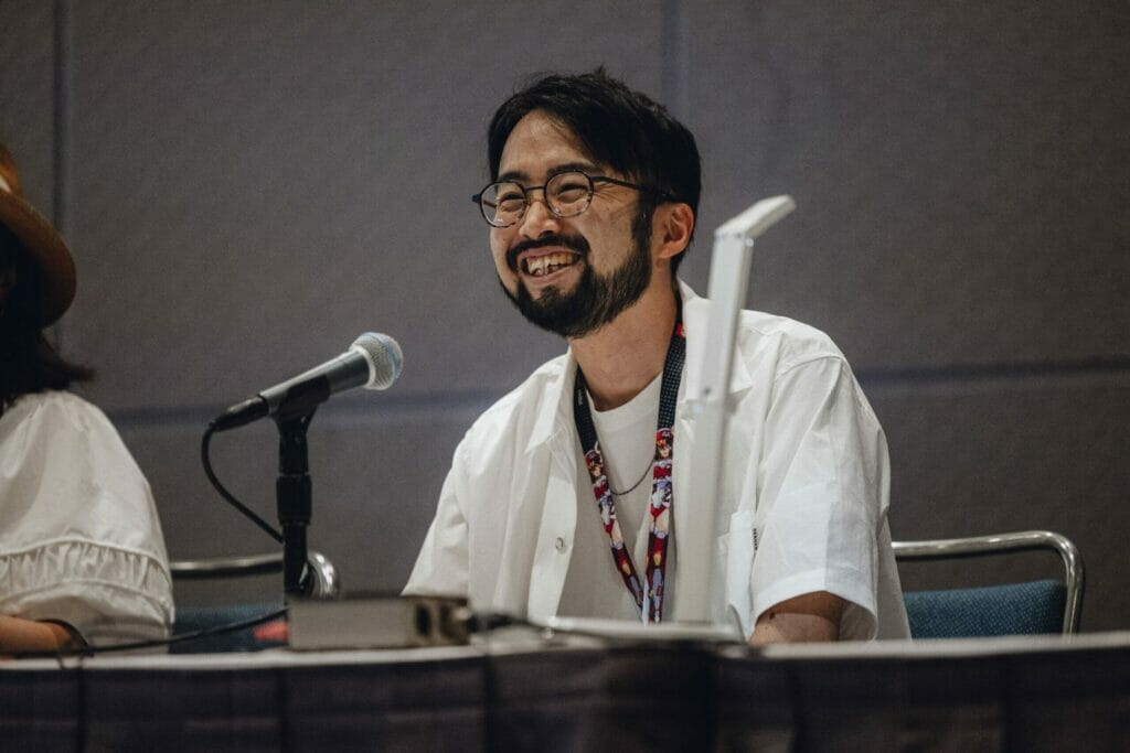 A member of the Ancient Magus' Bride anime staff smiles as he talks into a microphone.