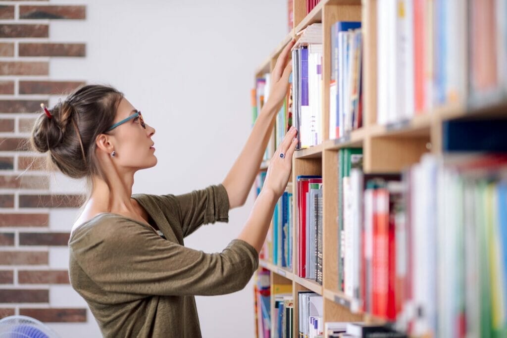 Photograph of a woman wearing glasses and cotton blouse looking for a book on a colorful bookshelf, in a library.