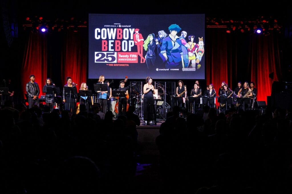 Photo of The Sinfionetta onstage at The Town Hall in front of a large screen, which features a visual for the Cowboy Bebop 25th Anniversary. Macy Schmidt stands in the center as the orchestra greets the audience.