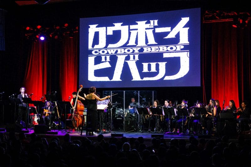 Photo of The Sinfionetta performing onstage at The Town Hall in front of a large screen, which features the Cowboy Bebop logo in black and white.