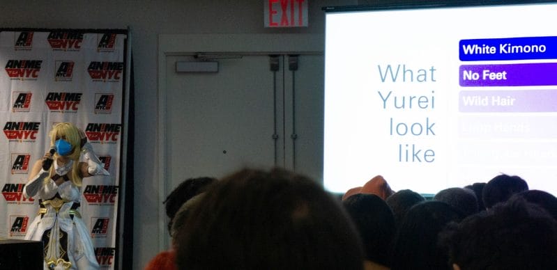 Conspiracy_Web, a woman dressed as Lumine from Genshin Impact, stands beside a PowerPoint slide that says "What Yurei Look Like"