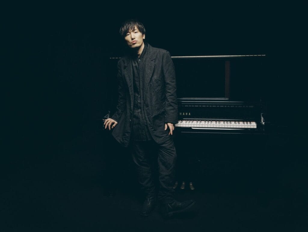 Hiroyuki Sawano is wearing a black suit, as he stands before a grand piano.