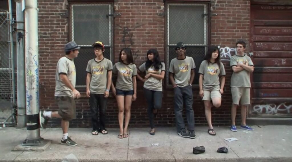 Screenshot from America's Greatest Otaku that depicts Stu Levey taling with the show's contestants, who are standing against a graffitied brick wall.