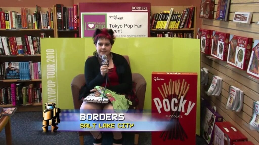 Screenshot from America's Greatest Otaku that depicts a pink-haired woman in a red top and black blazer talking into a microphone. She's sitting in a leather chair at a Borders bookstore, sitting in front of a green background and next to a giant box of Pocky.Chyron: "Borders: Salt Lake City"