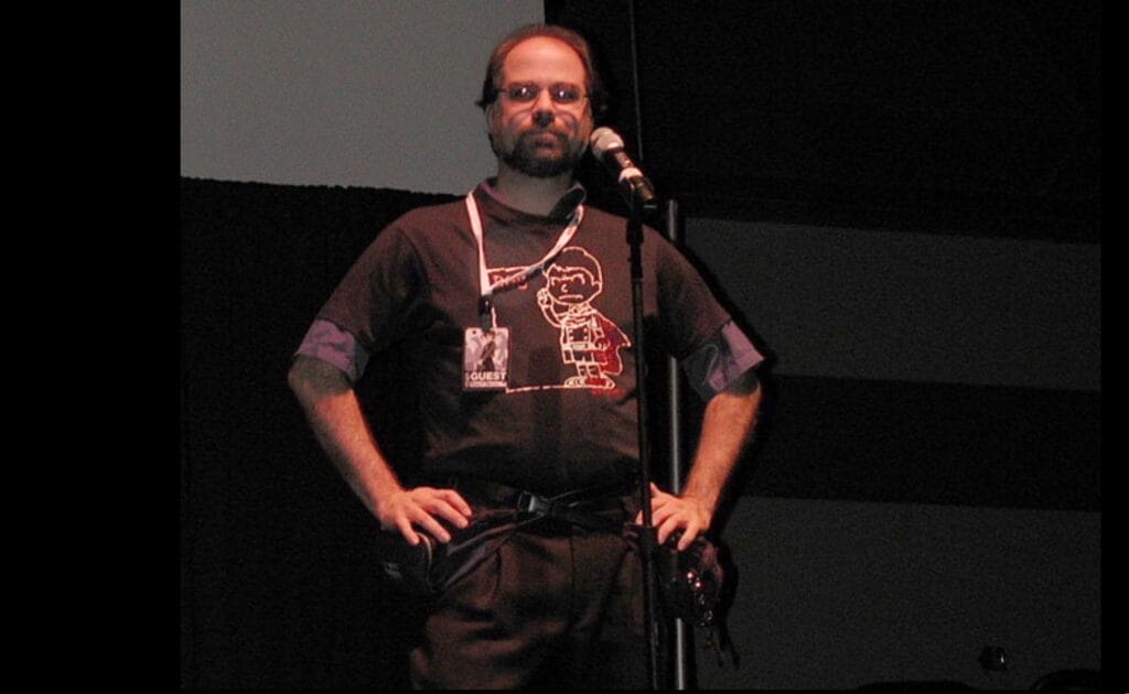 Robert Fenelon stands with his hands on his hips on the stage of a panel room at Otakon 2000