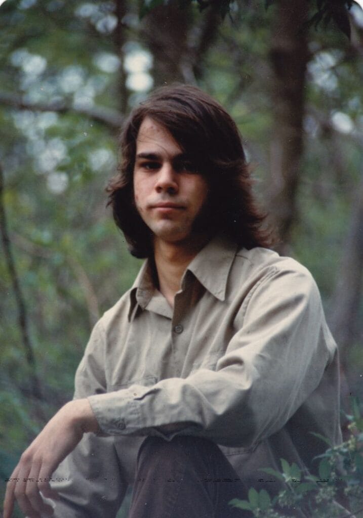 A photograph of Robert Fenelon, taken in the summer of 1981. He's young, with long brown hair and wearing a tan button-down shirt.