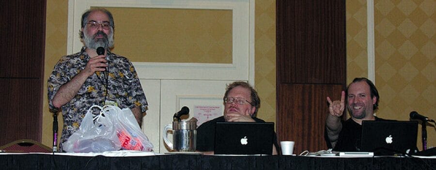 Photograph of three older men hosting a panel at a convention. Robert Fenelon is on the left, Walter Amos is in the middle, and Brian T. Price is on the right throwing the devil horns.