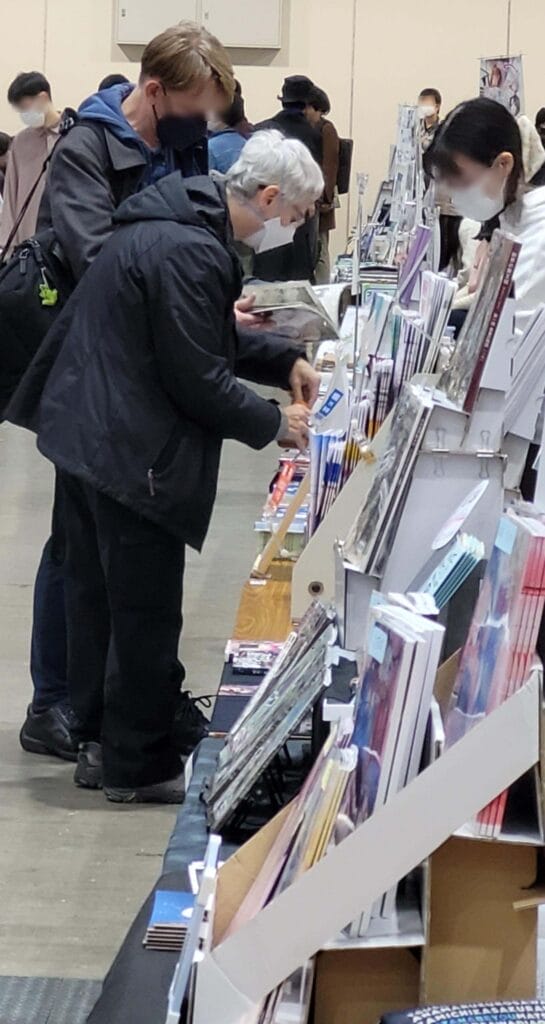 Erica Friedman at Comitia 146, looking through doujinshi as a shopkeeper minds her booth