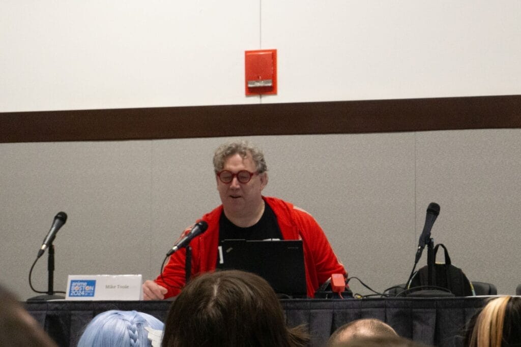 Photograph of Mike Toole at Anime Boston 2024. He's sitting behind a placard with his name on it, and reading from a laptop computer.