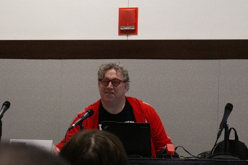 Photograph of Mike Toole at Anime Boston 2024. He's sitting behind a placard with his name on it, and reading from a laptop computer.