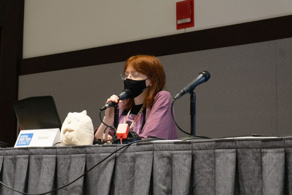 Photo of YouTuber Red Bard sitting behind a microphone, next to a plushie. A placard with their name sits nearby.