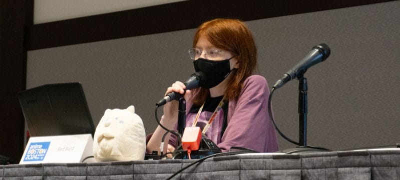 Photo of YouTuber Red Bard sitting behind a microphone, next to a plushie. A placard with their name sits nearby.