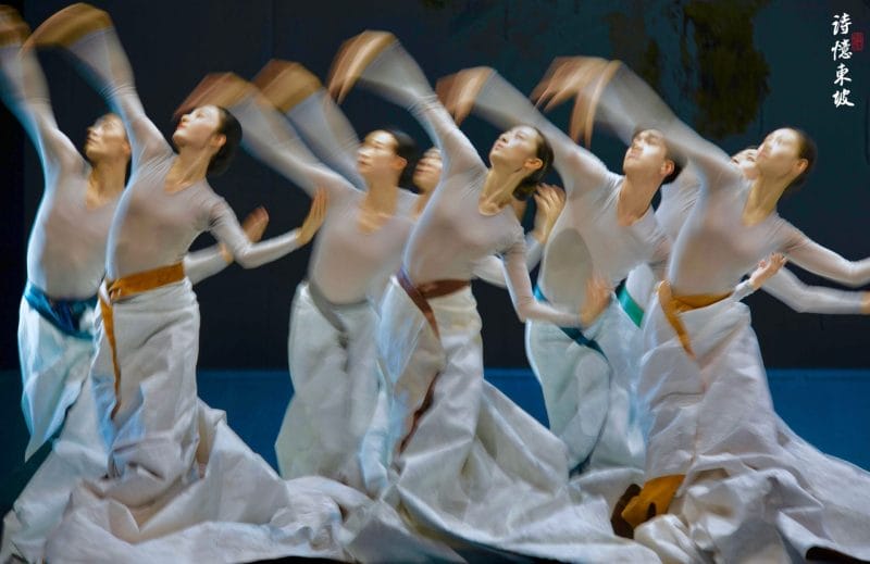 Photograph from Dongpo: Life In Poems that depicts dancers in grey tops and blue skirts performing against a black backdrop.