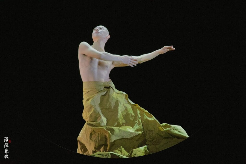 Photograph from Dongpo: Life In Poems that depicts a masculine, bald dancer, wearing a yellow skirt