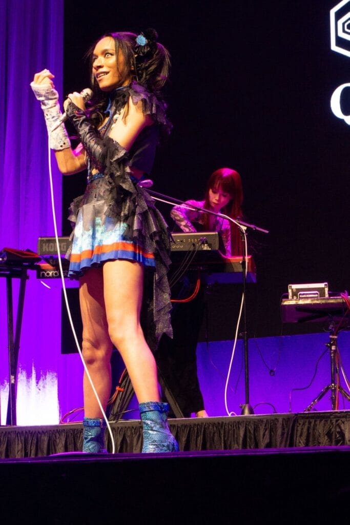 Photograph of Queen Bee performing at Anime Boston 2024. Avu-chan is dressed in a black, blue, and red outfit with sky blue boots, and standing with a mischievous smile.
