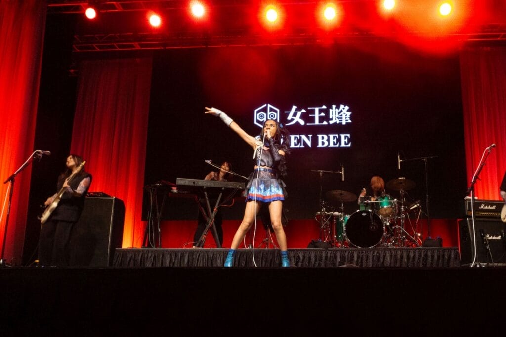 Photograph of Queen Bee performing at Anime Boston 2024. Avu-chan is dressed in a black, blue, and red outfit with sky blue boots, as Yashi-chan plays bass stage right.