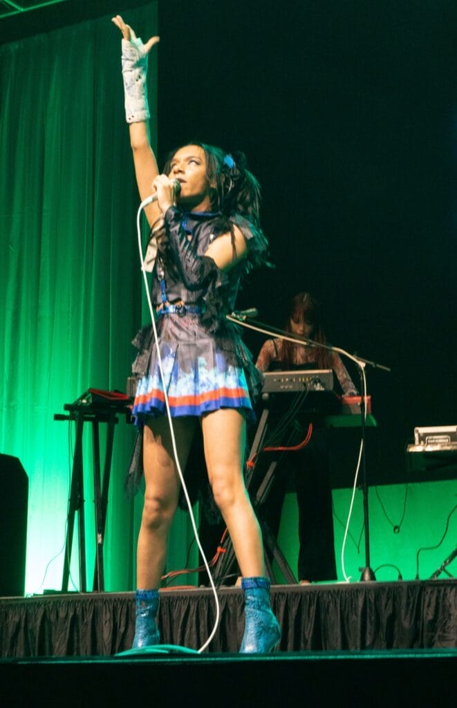 Photograph of Queen Bee performing at Anime Boston 2024. Avu-chan is dressed in a black, blue, and red outfit with sky blue boots, reaching up as she sings