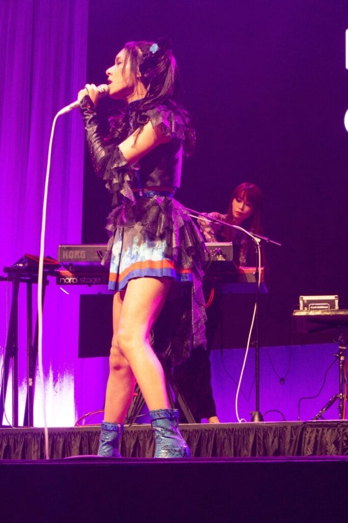 Photograph of Queen Bee performing at Anime Boston 2024. Avu-chan is dressed in a black, blue, and red outfit with sky blue boots, standing straight as she sings.