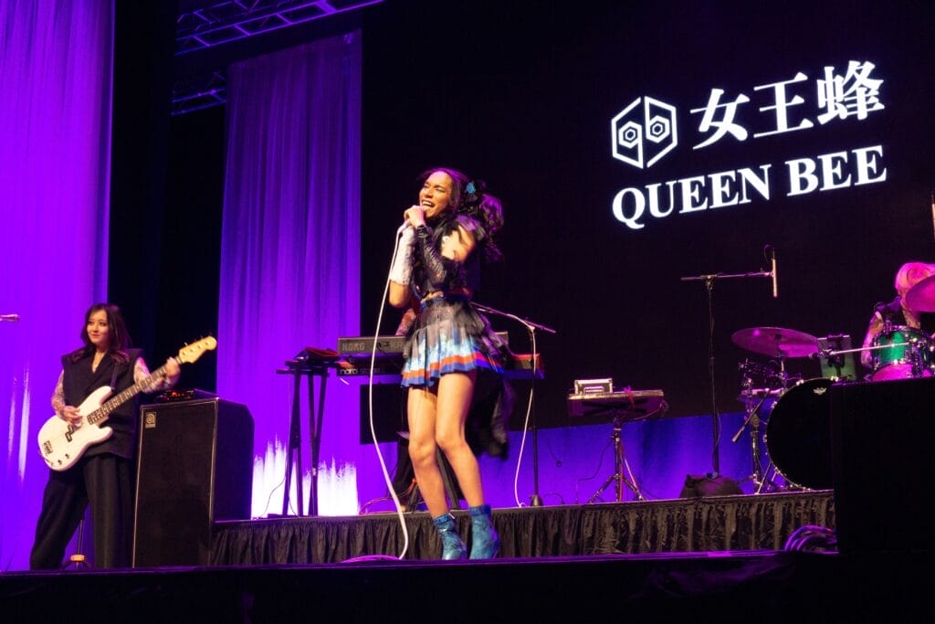 Photograph of Queen Bee performing at Anime Boston 2024. Avu-chan is dressed in a black, blue, and red outfit with sky blue boots while Yashi-chan plays bass wearing a sleeveless black shirt.