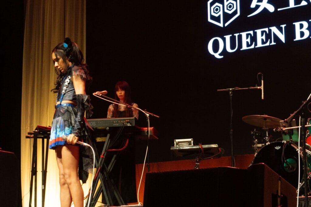 Photograph of Queen Bee performing at Anime Boston 2024. Avu-chan is dressed in a black, blue, and red outfit with sky blue boots, standing straight and looking down with a sad expression