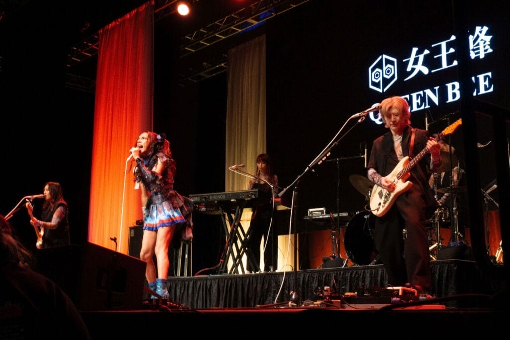 Photograph of Queen Bee performing at Anime Boston 2024. Avu-chan is dressed in a black, blue, and red outfit with sky blue boots, standing straight as they sing. Yashi-chan, meanwhile, plays the bass wearing a black sleeveless shirt