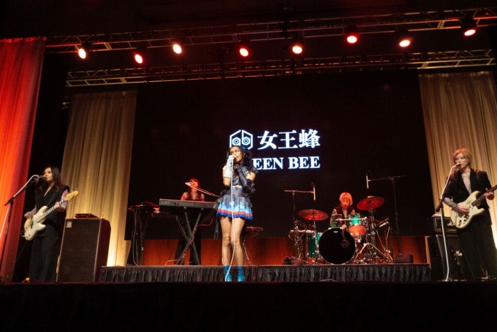 Photograph of Queen Bee performing at Anime Boston 2024. Avu-chan is dressed in a black, blue, and red outfit with sky blue boots, standing straight as they sing. Hibari-kun, meanwhile, plays the guitar while wearing a black, short-sleeved shirt and Yashi-chan plays bass  wearing a black sleeveless shirt