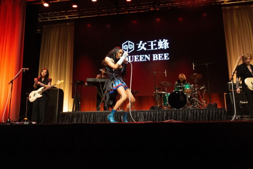 Photograph of Queen Bee performing at Anime Boston 2024. Avu-chan is dressed in a black, blue, and red outfit with sky blue boots while Yashi-chan plays bass wearing a sleeveless black shirt. Hibari-kun can be seen stage left, wearing a black shirt and slacks.