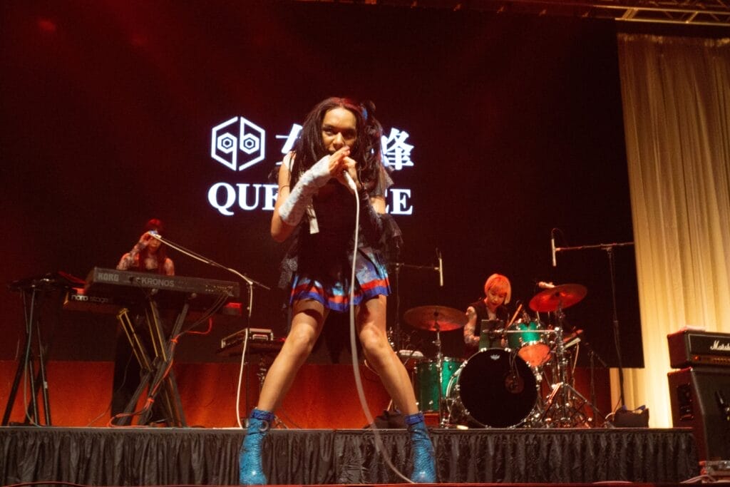Photograph of Queen Bee performing at Anime Boston 2024. Avu-chan is dressed in a black, blue, and red outfit with sky blue boots. She's leaning into the camera as she sings.
