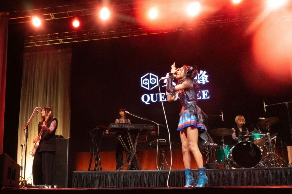 Photograph of Queen Bee performing at Anime Boston 2024. Avu-chan is dressed in a black, blue, and red outfit with sky blue boots, standing straight as they sing. Yashi-chan, meanwhile, plays the bass wearing a black sleeveless shirt