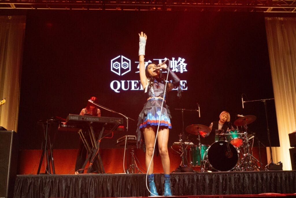 Photograph of Queen Bee performing at Anime Boston 2024. Avu-chan is dressed in a black, blue, and red outfit with sky blue boots, reaching up as she leans back and belts out a note.